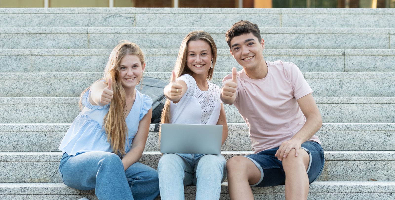 Drei Jugendliche sitzen auf einer Treppe und recken lachend ihren Daumen in die Kamera
