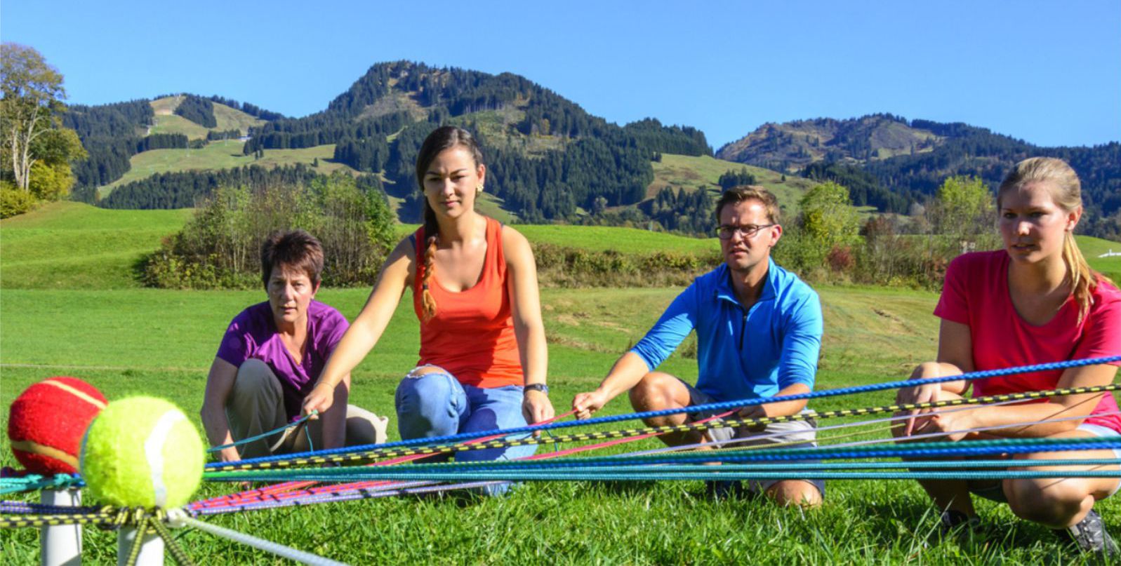 Eine Gruppe Erwachsener sitzt im Freien im Kreis und versucht mittels Schnren Blle zu dirigieren